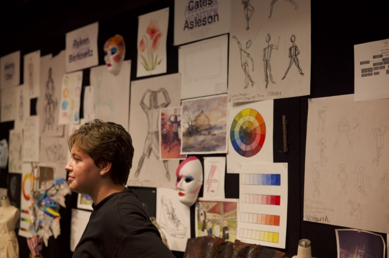 Drama: Production & Design student stands in profile with their final project, which includes sketches pinned to the wall and a face mask.